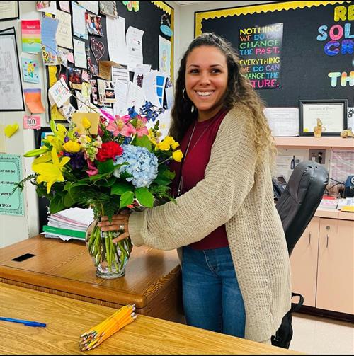 Mrs. Nayelli standing at her desk with a bouquet of flowers in a vase.